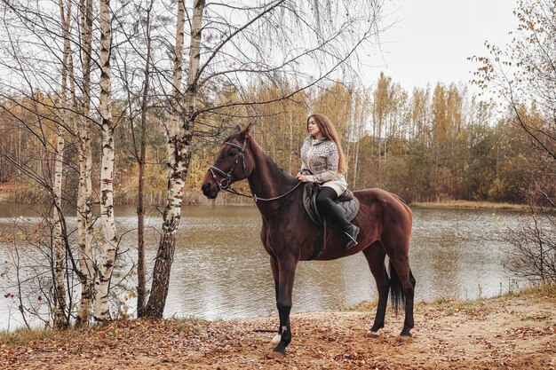 Mulher jovem e bonita a cavalo na floresta de outono pelo lago