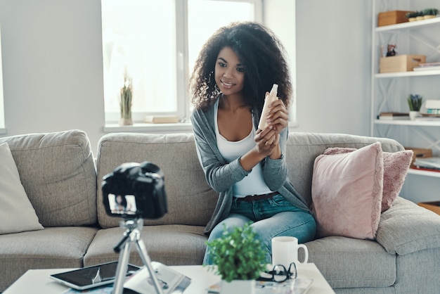 Mulher jovem e bonita africana apontando para um produto de beleza e sorrindo enquanto faz um vídeo nas redes sociais