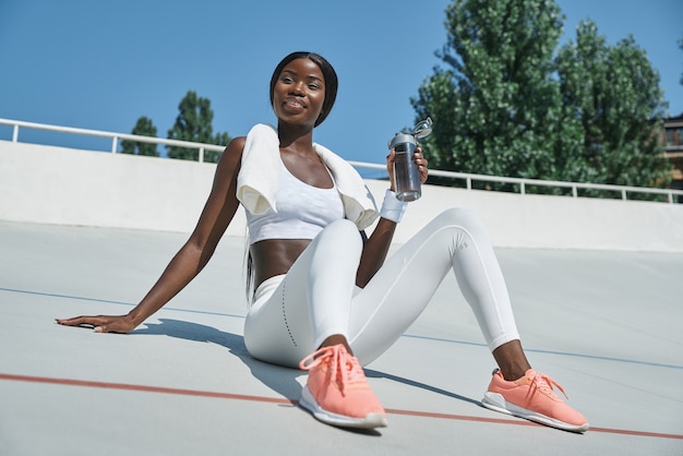 Mulher jovem e bonita africana em roupas esportivas, segurando a garrafa com água enquanto está sentado no esporte t.