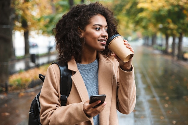 Mulher jovem e bonita africana vestindo um casaco, caminhando ao ar livre no parque, carregando uma mochila, usando o telefone celular, bebendo café