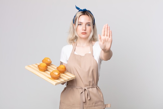 Mulher jovem e bonita albina olhando séria, mostrando a palma da mão aberta, fazendo um gesto de pare com uma bandeja de muffins