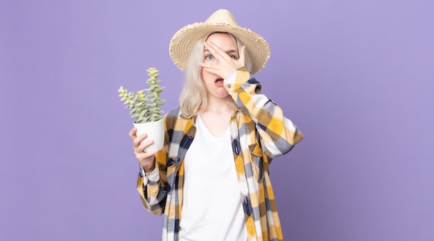 Mulher jovem e bonita albina parecendo chocada, assustada ou apavorada, cobrindo o rosto com a mão e segurando um cacto de planta doméstica