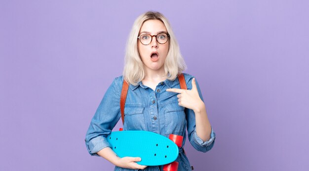 Mulher jovem e bonita albina parecendo chocada e surpresa com a boca bem aberta, apontando para si mesma e segurando um skate