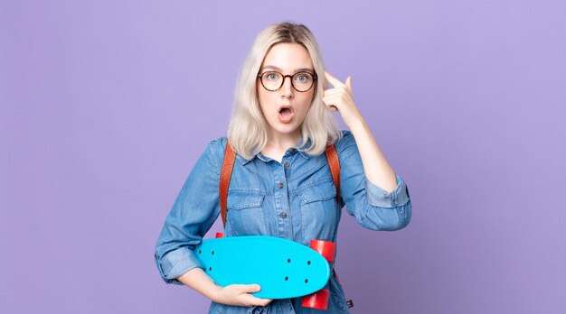 Mulher jovem e bonita albina parecendo surpresa, percebendo um novo pensamento, ideia ou conceito e segurando um skate