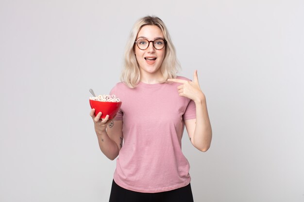 Mulher jovem e bonita albina se sentindo feliz e apontando para si mesma com um animado com uma tigela de café da manhã