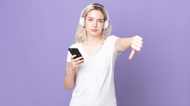 Mulher jovem e bonita albina se sentindo mal, mostrando os polegares para baixo com fones de ouvido e smartphone