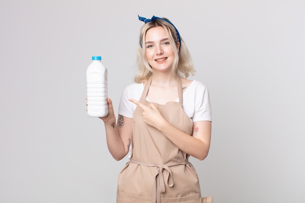 Mulher jovem e bonita albina sorrindo alegremente, se sentindo feliz e apontando para o lado com uma garrafa de leite