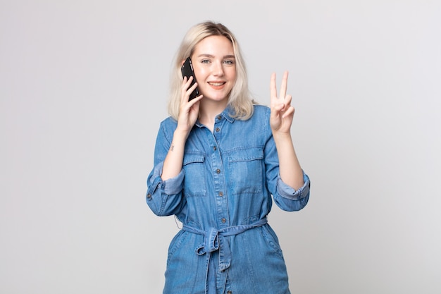 Mulher jovem e bonita albina sorrindo e parecendo amistosa, mostrando o número dois e falando com um smartphone