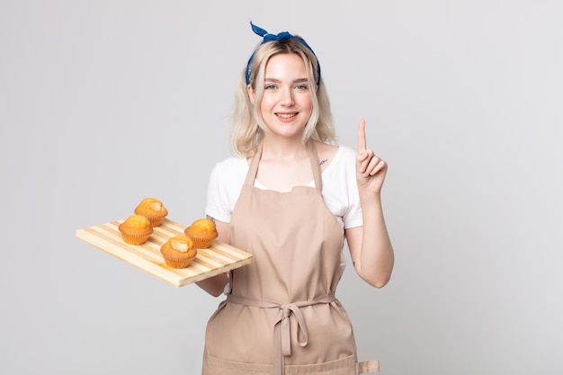 Mulher jovem e bonita albina sorrindo e parecendo amistosa, mostrando o número um com uma bandeja de muffins