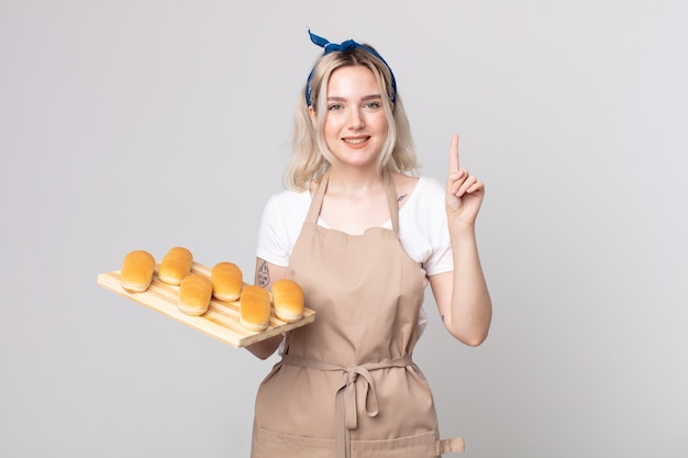 Mulher jovem e bonita albina sorrindo e parecendo amistosa, mostrando o número um com uma bandeja de pães