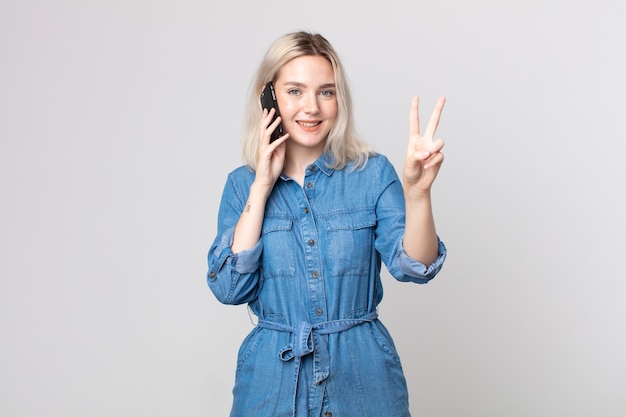 Mulher jovem e bonita albina sorrindo e parecendo feliz, gesticulando vitória ou paz e falando com um smartphone