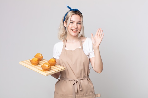 Mulher jovem e bonita albina sorrindo feliz, acenando com a mão, dando as boas-vindas e cumprimentando você com uma bandeja de muffins