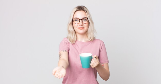 Mulher jovem e bonita albina sorrindo feliz com simpatia e oferecendo e mostrando um conceito com uma caneca de café