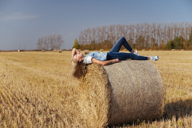 Mulher jovem e bonita aldeã posando de jeans em um fardo de feno em um campo
