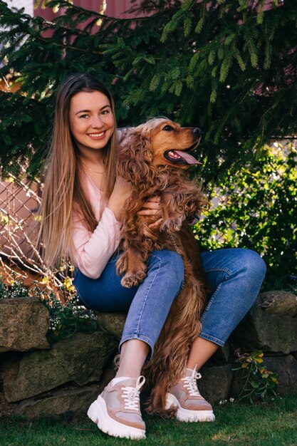 Mulher jovem e bonita alegre com cabelo comprido, sentado e abraçando o cachorro no quintal perto da árvore.