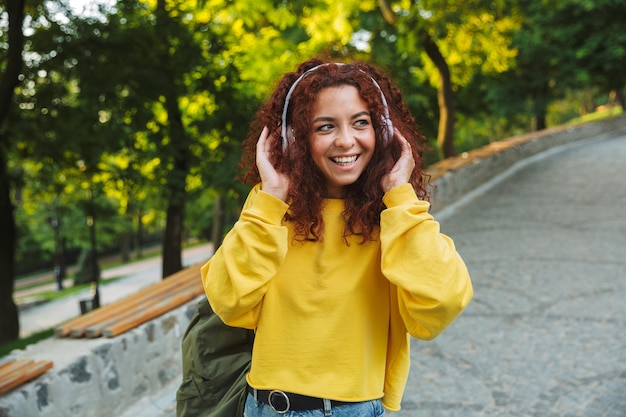 Mulher jovem e bonita alegre passando um bom tempo no parque, ouvindo música com fones de ouvido enquanto caminha