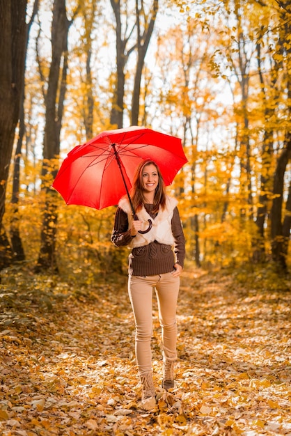 Mulher jovem e bonita andando com guarda-chuva vermelho pela floresta ensolarada em cores de outono.
