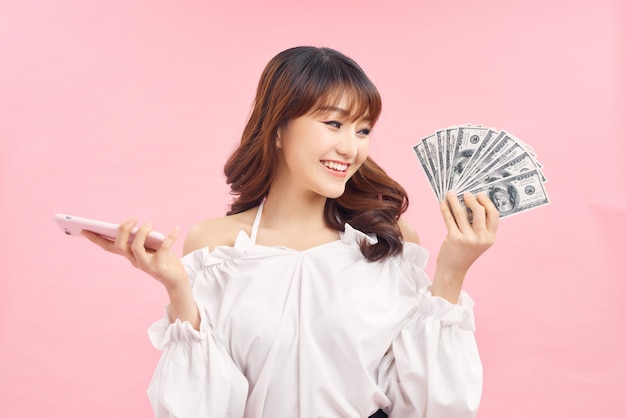 mulher jovem e bonita animada posando isolado sobre o fundo da parede rosa, segurando dinheiro e usando telefone celular.