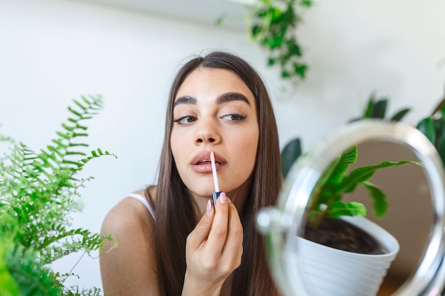 Mulher jovem e bonita aplicando brilho labial com aplicador na frente de um espelho em casa