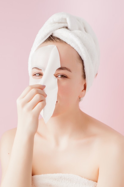 Mulher jovem e bonita aplicando uma máscara de tecido cosmético em um rosto em rosa