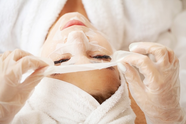 Foto mulher jovem e bonita aplicando uma máscara de tecido cosmético em um rosto
