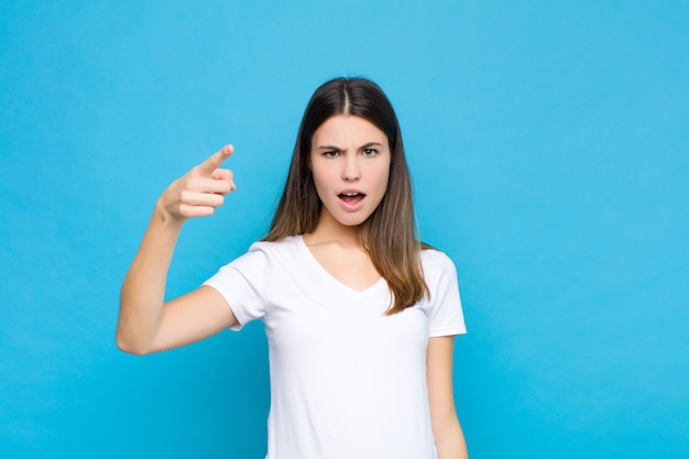 Mulher jovem e bonita apontando com uma expressão agressiva e zangada, parecendo um chefe louco e furioso contra uma parede azul