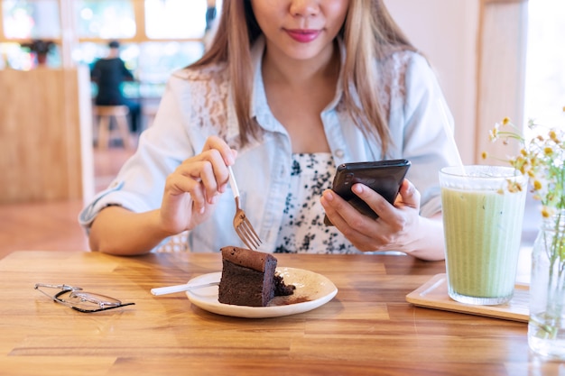 Mulher jovem e bonita asiática comendo bolo de chocolate enquanto usa o smartphone