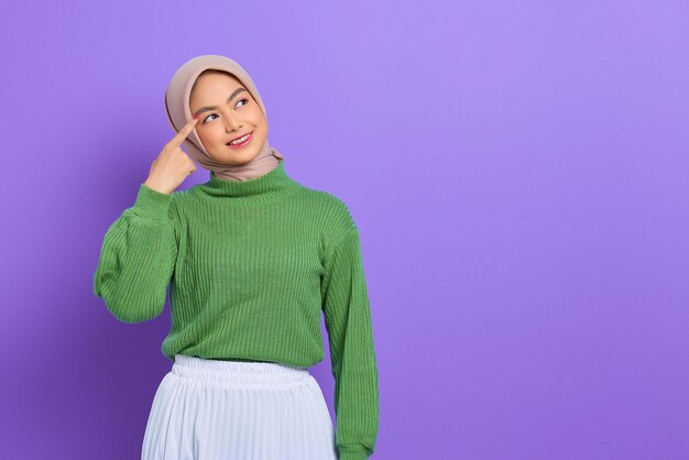 Mulher jovem e bonita asiática em suéter verde sorrindo, olhando para longe com um dedo na cabeça, tendo uma ótima ideia isolada sobre fundo roxo