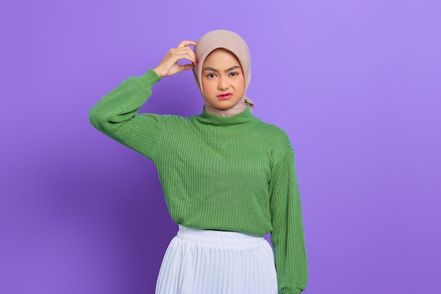Mulher jovem e bonita asiática em um suéter verde confusa com uma pergunta, pensando com a mão na cabeça isolada sobre um fundo roxo