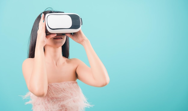 Mulher jovem e bonita asiática feliz em pé, sorrindo para ela usando um fone de ouvido de realidade virtual, jogando em um dispositivo de óculos de realidade virtual em um fundo azul com espaço de cópia para o texto