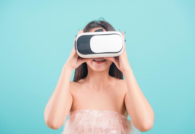 Mulher jovem e bonita asiática feliz em pé, sorrindo para ela usando um fone de ouvido de realidade virtual, jogando em um dispositivo de óculos de realidade virtual em um fundo azul com espaço de cópia para o texto
