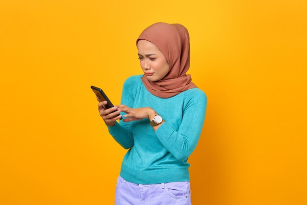 Mulher jovem e bonita asiática triste usando smartphone isolado em fundo amarelo