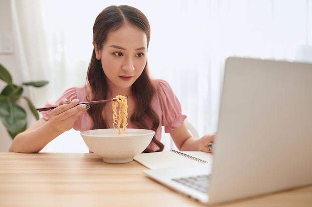 Mulher jovem e bonita asiática usando laptop e comendo macarrão asiático em casa.