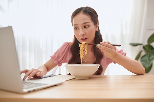 Mulher jovem e bonita asiática usando laptop e comendo macarrão asiático em casa.