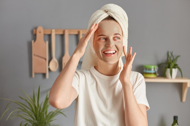 Mulher jovem e bonita atraente posando na cozinha com toalha na cabeça vestindo camiseta branca casual sorrindo olhando para longe desfrutando de cuidados com a pele anti envelhecimento procedimentos hidratantes