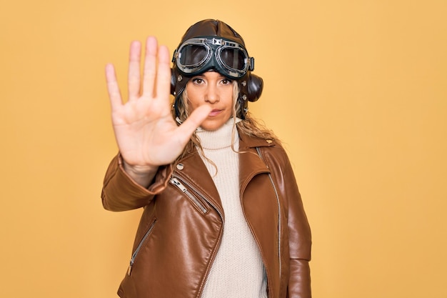 Mulher jovem e bonita aviadora loira usando capacete de piloto vintage com óculos e jaqueta parando de cantar com a palma da mão Expressão de advertência com gesto negativo e sério no rosto