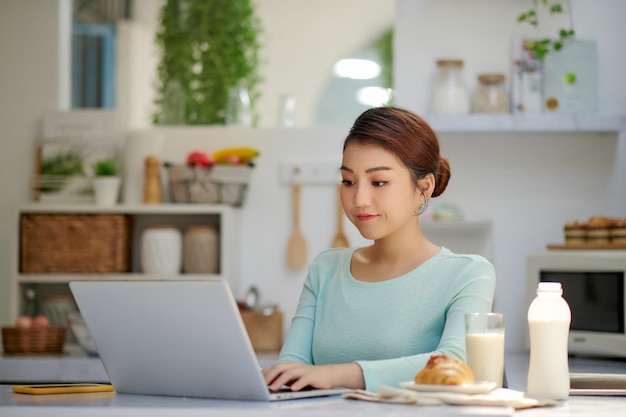 Mulher jovem e bonita bebendo smoothie delicioso saudável e usando laptop em casa