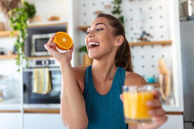 Mulher jovem e bonita bebendo suco de laranja fresco na cozinha dieta saudável mulher jovem feliz com copo de suco e laranja na mesa na cozinha