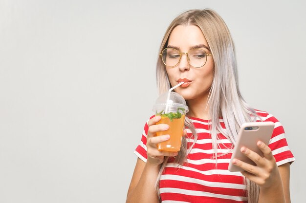 Mulher jovem e bonita bebendo um suco de desintoxicação caseiro, enviando mensagens de texto em seu telefone enquanto isolado sobre o branco. Usando o telefone.