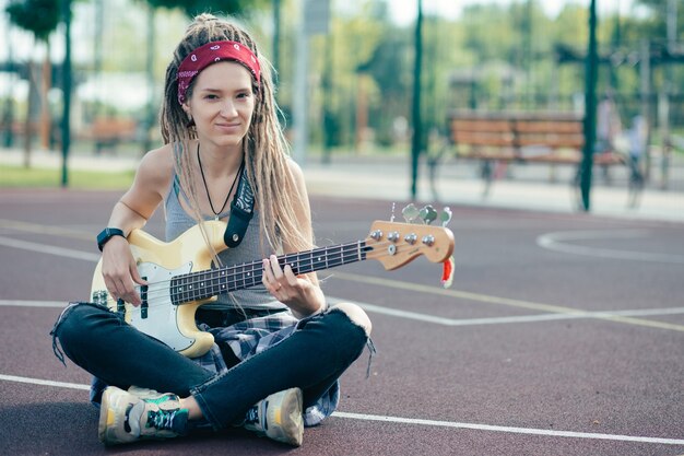 Mulher jovem e bonita calma com dreadlocks sentada no chão de um campo de esportes e olhando para você enquanto segura seu violão