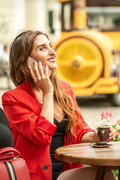 Mulher jovem e bonita caucasiana feliz falando ao celular, sentada em um café ao ar livre em uma cidade europeia