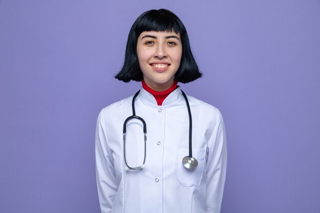 Mulher jovem e bonita caucasiana sorridente com uniforme de médico e estetoscópio olhando