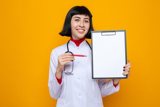 Mulher jovem e bonita caucasiana sorridente com uniforme de médico e estetoscópio segurando uma caneta e uma prancheta