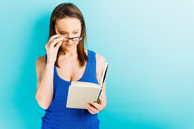 Mulher jovem e bonita colocando os óculos enquanto tenta ler um livro sobre fundo azul