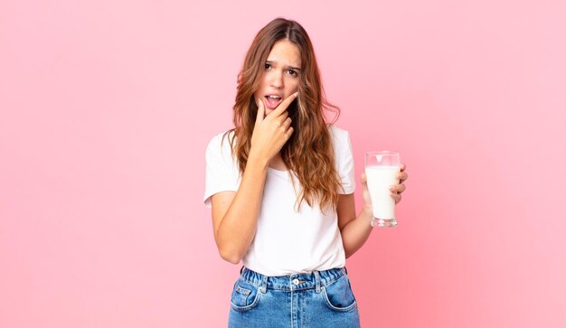 Mulher jovem e bonita com a boca e os olhos bem abertos, a mão no queixo e segurando um copo de leite