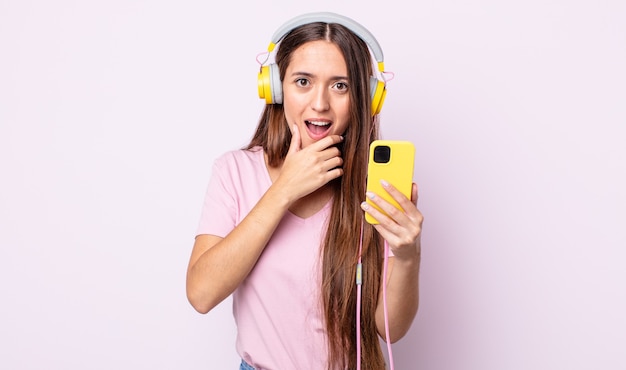 Mulher jovem e bonita com a boca e os olhos bem abertos e a mão no queixo. fones de ouvido e smartphone