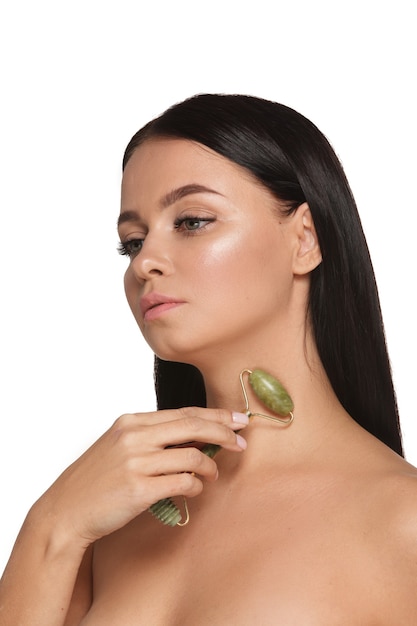 Mulher jovem e bonita com a pele perfeita usando uma toalha na cabeça usando um rolo facial de jade com pedras naturais de quartzo em branco