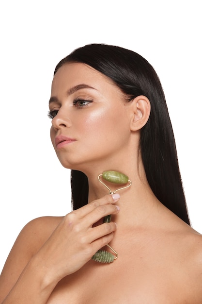 Mulher jovem e bonita com a pele perfeita usando uma toalha na cabeça usando um rolo facial de jade com pedras naturais de quartzo em branco