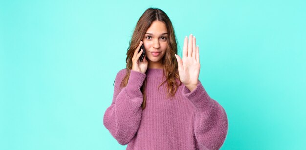 Mulher jovem e bonita com aparência séria, mostrando a palma da mão aberta, fazendo gesto de parada e usando um smartphone