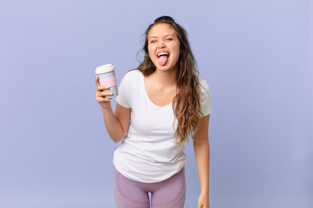 Foto mulher jovem e bonita com atitude alegre e rebelde, brincando e mostrando a língua e segurando um café